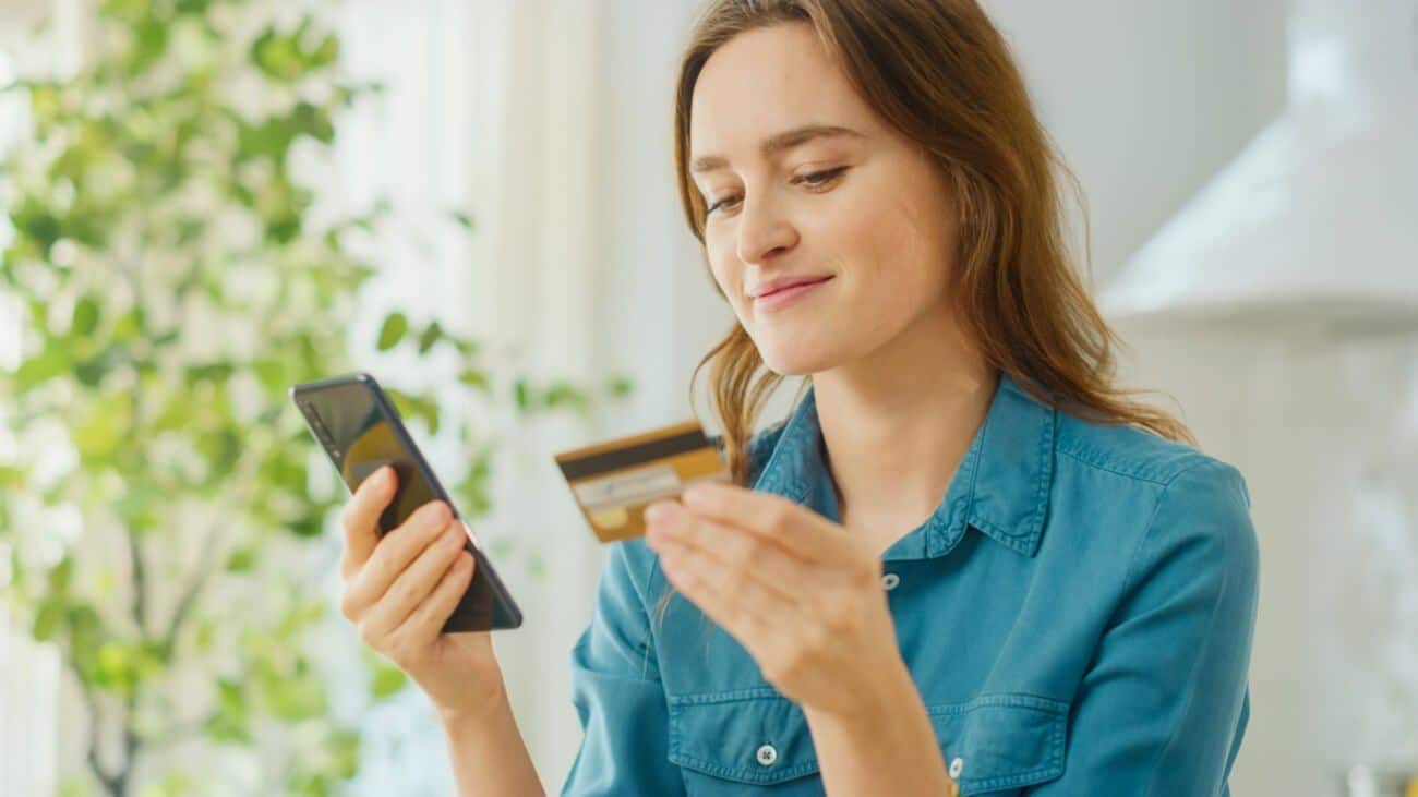 woman with brown hair and blue short looking at a credit card and typing into her phone