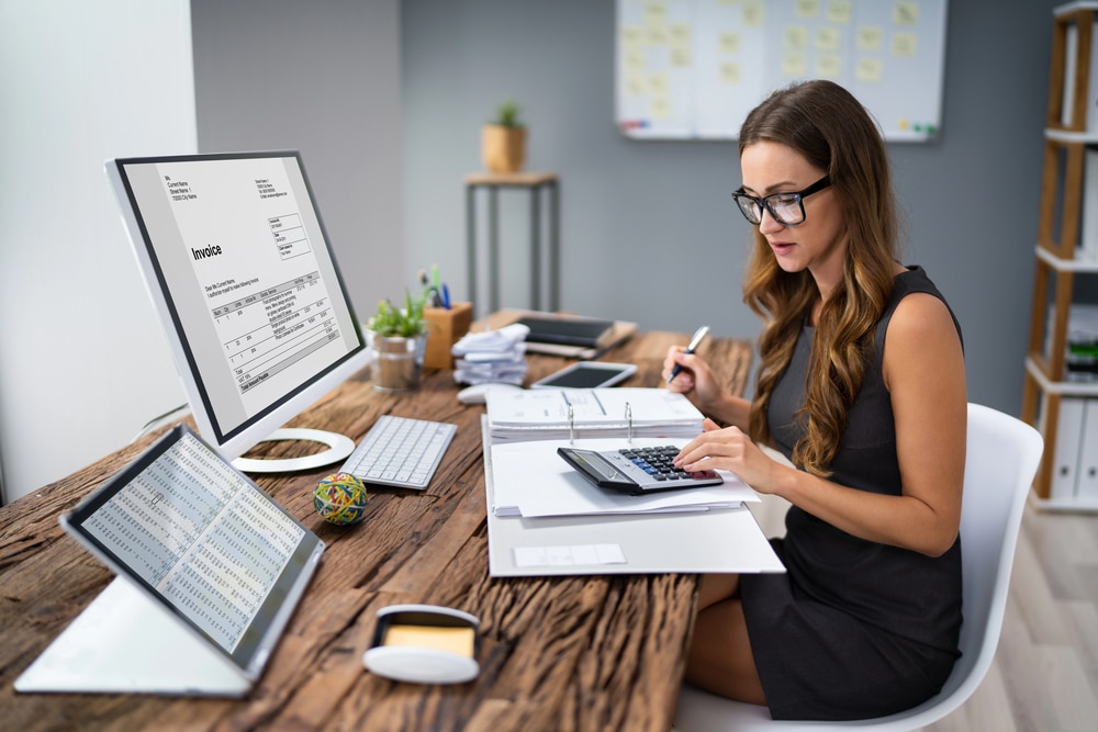 woman wearing black dress and glasses sitting at a wood desk holding a pen and using a calculator on top of a binder with papers